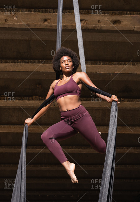 Slender African American female balancing on hanging hammock while doing aerial yoga above ground under bridge in city