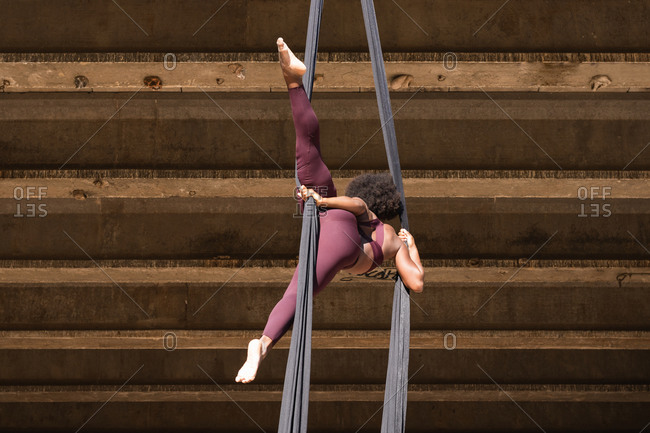 Slender African American female balancing on hanging hammock while doing aerial yoga above ground under bridge in city