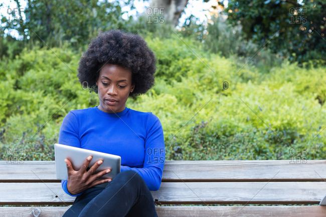 African American female freelancer sitting on bench in park and using tablet while working remotely on startup project