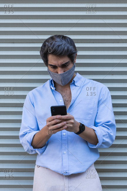 Serious male in stylish clothes and protective mask standing near building in city and texting messages on cellphone during coronavirus outbreak