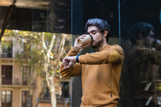 Side view of male checking time on wristwatch and drinking coffee to go while standing on street and waiting for appointment