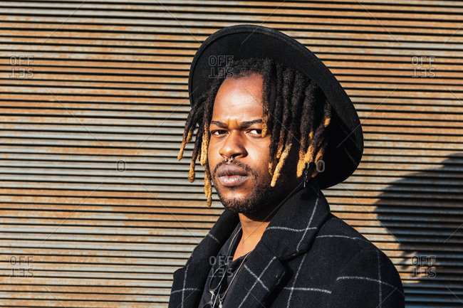 Modern adult black man with dreadlocks wearing hat and stylish coat looking at camera against street wall in sunlight