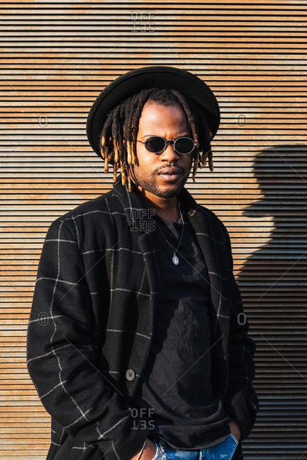 Modern adult black man with dreadlocks wearing hat, sunglasses and stylish coat looking at camera against street wall in sunlight