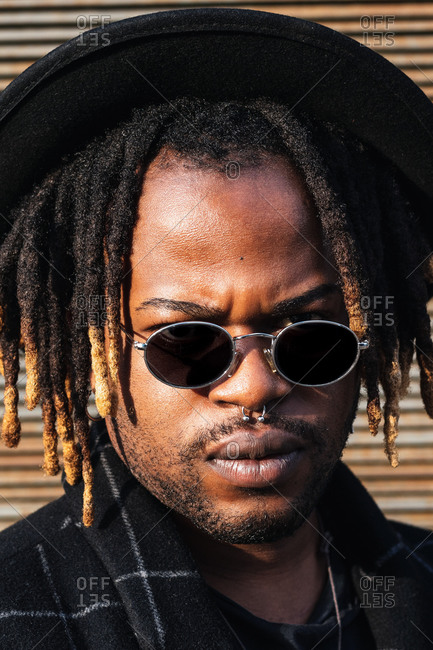 Modern adult black man with dreadlocks wearing hat, sunglasses and stylish coat looking at camera against street wall in sunlight stock photo - OFFSET Modern adult black man with dreadlocks wearing hat, sunglasses and stylish coat looking at camera against street wall in sunlight stock photo - OFFSET