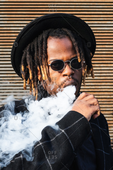 Modern African American man with dreadlocks wearing hat with sunglasses and exhaling smoke on street