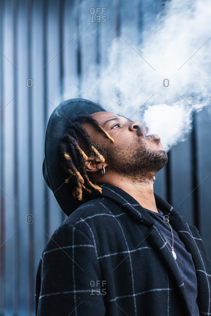 Side view of ethnic back guy with dreadlocks and hat in dense cloud of fume smoking on street in city