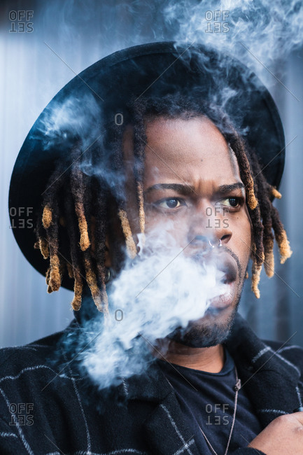 Side view of ethnic back guy with dreadlocks and hat in dense cloud of fume smoking on street in city