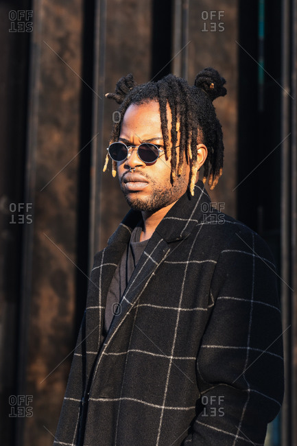 Frowning modern African American guy with dreadlocks hairstyle wearing sunglasses with coat standing in sunlight