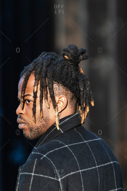 Side view of frowning modern African American guy with dreadlocks hairstyle wearing coat standing on the street looking away