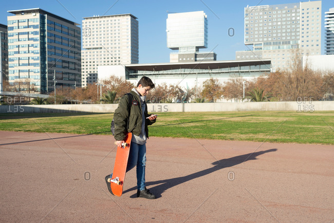 Full body side view of contemporary male teenager in warm jacket carrying skateboard and using mobile phone while walking on city street