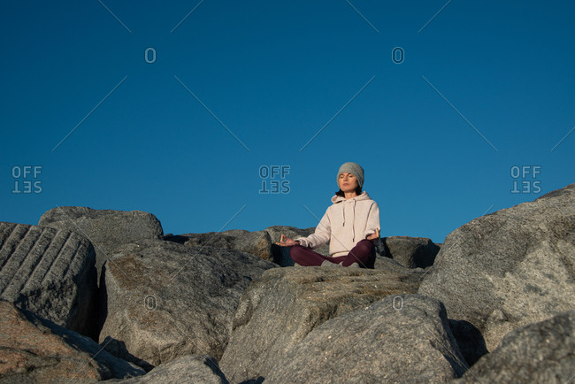 Young female in warm activewear and knitted hat meditating with mudra hand gesture while sitting on rock against blue sky