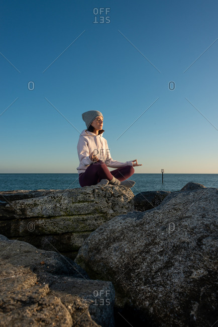 Side view of young female in warm activewear and knitted hat meditating with mudra hand gesture while sitting on rock against blue sky