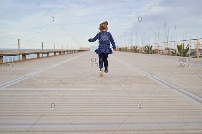 Back view of adorable cheerful little girl in blue dress with toy on hand having fun on paved walkway in summer day