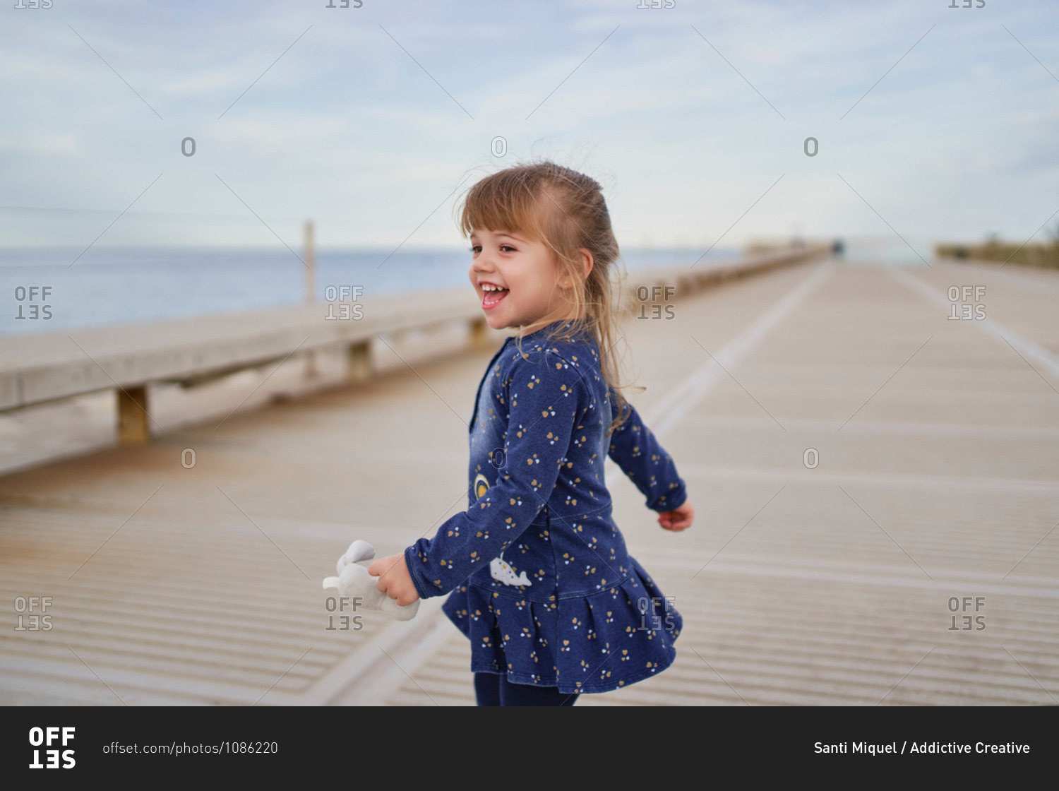 High angle of adorable cheerful little girl in blue dress with toy on hand  having fun on paved walkway in summer day stock photo - OFFSET