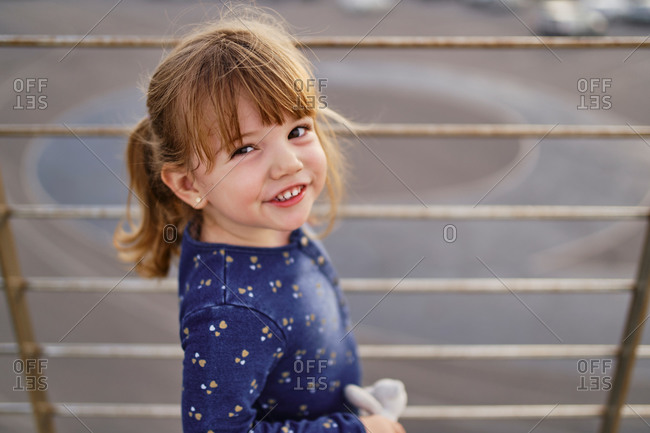 High angle of adorable cheerful little girl in blue dress with toy on hand having fun on paved walkway in summer day