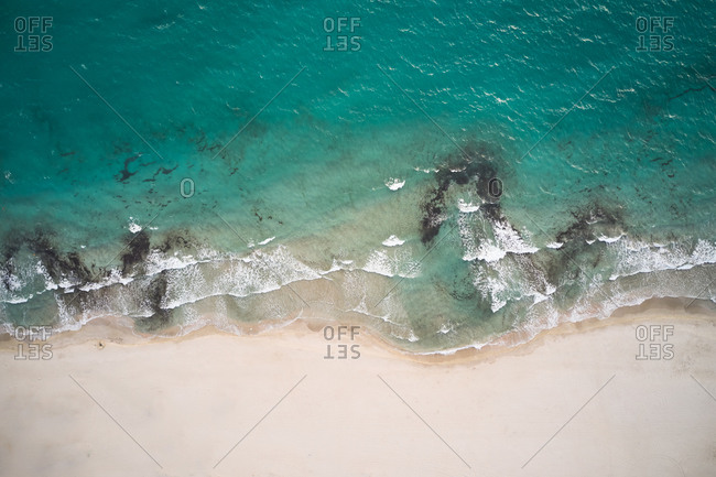 Aerial top view of turquoise water with seaweed and foamy waves rolling on empty sandy beach
