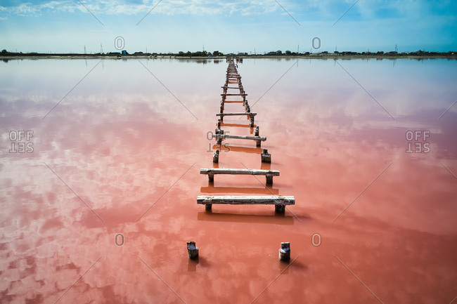 Drone view of old wooden walkway leading through pink water of calm lake towards distant coast