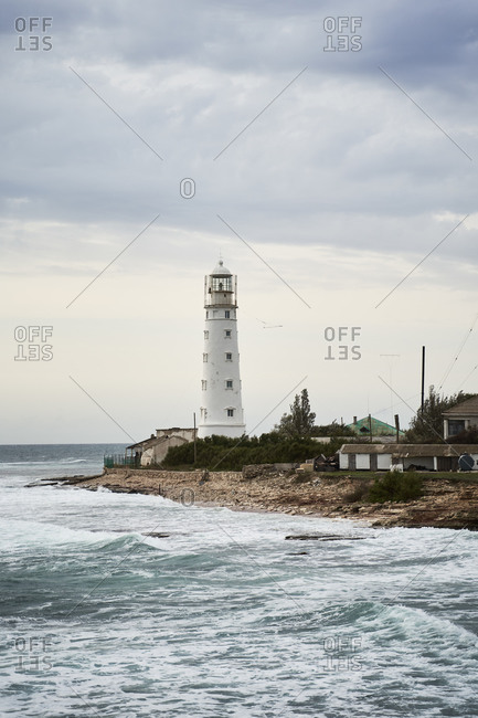 Tower of white beacon located on coast near waving sea under overcast sky