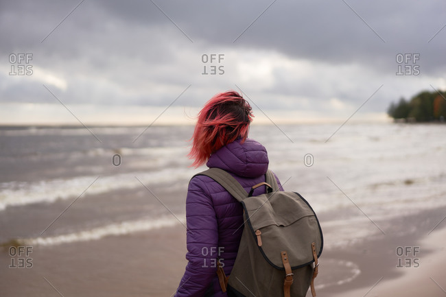 Back view of unrecognizable female traveler in outerwear standing on shore and admiring calm sea on cloudy day in Sestroretsk in autumn