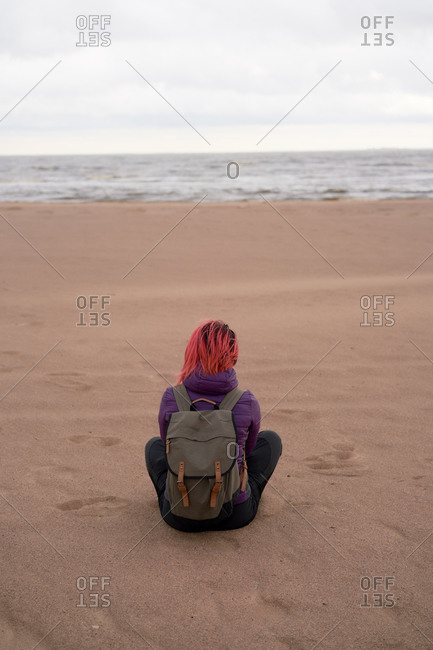 Back view of unrecognizable female traveler in outerwear siting on shore and admiring calm sea on cloudy day in Sestroretsk in autumn