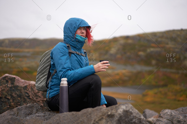 Side view of traveling woman in outerwear sitting on rock and enjoying hot tea from thermos cup while admiring mountainous landscape in autumn