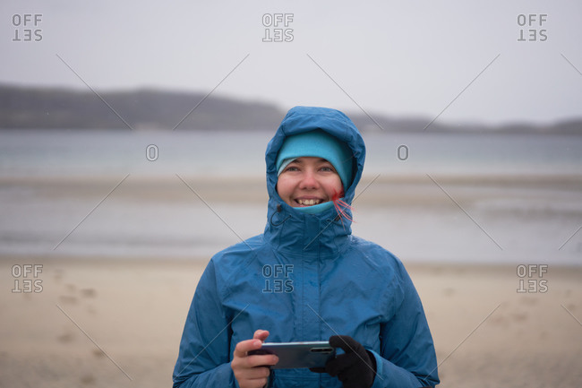 Delighted female explorer in warm waterproof jacket standing with cellphone at seaside on windy day and looking at camera