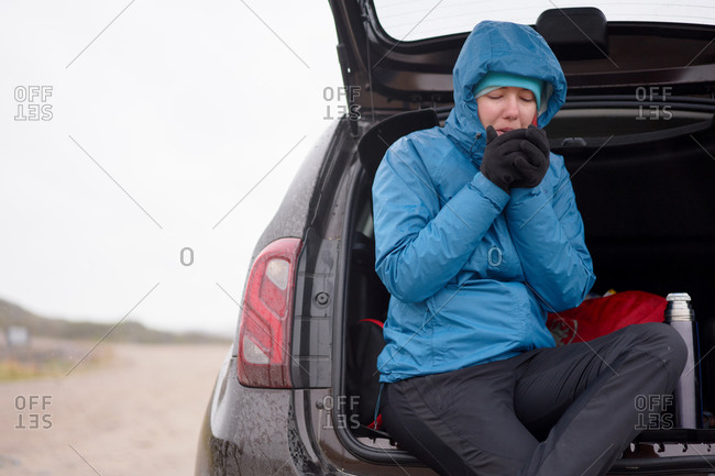 Female traveler in outerwear sitting in car trunk and warming hands with breath