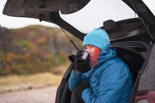 Side view of carefree female traveler in outerwear sitting in car trunk and enjoying hot drink from thermos cup while looking away