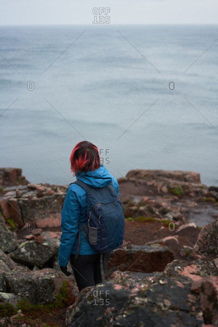 From above back view of unrecognizable explorer with backpack standing on rocky hill and observing amazing view of sea on cloudy day in autumn