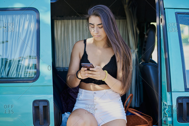 Young female in summer outfit sitting in van and reading messages on social media on smartphone