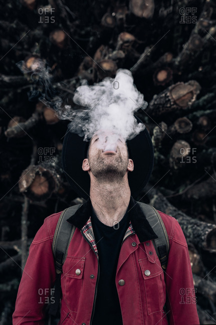 Male hipster in hat and red shirt exhaling cloud of smoke standing along with eyes closed against pile of logs in nature