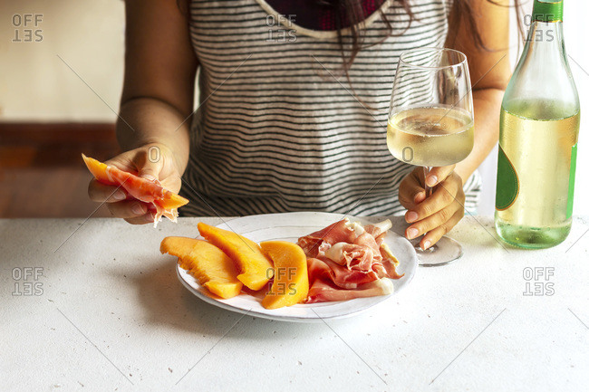 Female hands holding a sliced of cantaloupe wrapped in prosciutto and a glass of wine