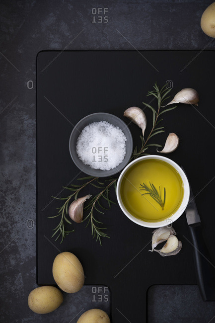 A bowl of olive oil, salt flakes, rosemary and potatoes on a black wooden cutting board.