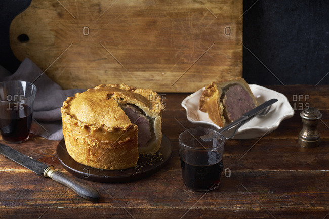 Traditional British raised pork pie with bacon and herbs with one slice cut and served next to a glass of red wine
