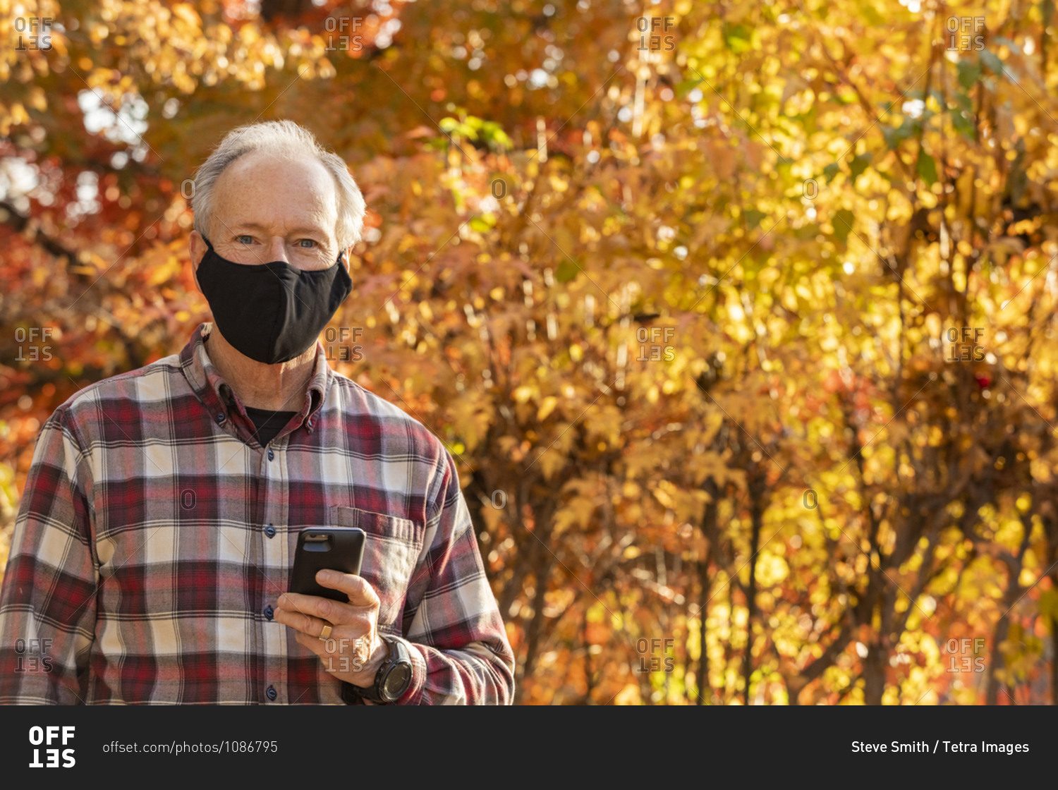 Senior man wearing Covid protective mask and holding smartphone outdoors in fall