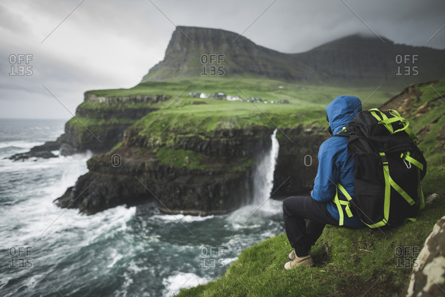 Denmark, Faroe Islands, Gasadalur Village, Mt. Lafossur Waterfall, Man with backpack sitting on edge of cliff and looking at Mulafossur Waterfall