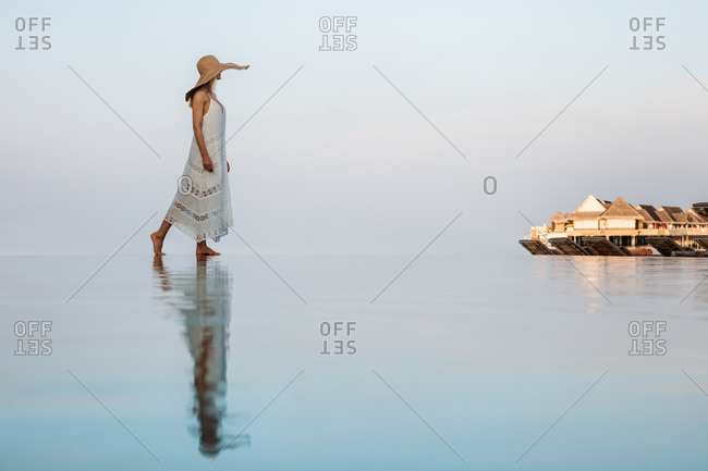 Side view of a woman in hat walking near an infinity pool with her reflection falling on water. Woman enjoying the scenic view of overwater villas at a luxury resort.