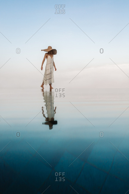 Scenic view of a woman walking at an infinity pool appearing to walk on water. Portrait of a woman walking at an infinity pool with her reflection falling on water.