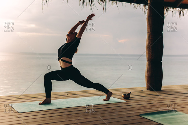 Woman standing on an overwater villa practicing yoga. Woman in warrior yoga position near the sea.
