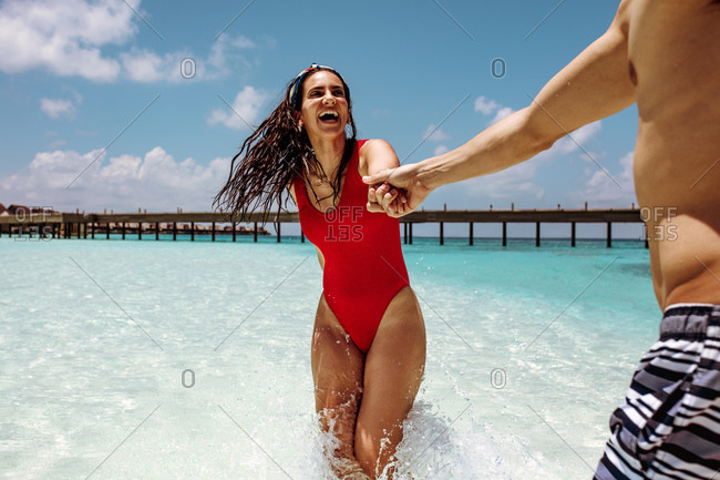 Tourist couple having fun on a tropical beach. Happy woman in swimsuit playing in water holding the hand of her partner.