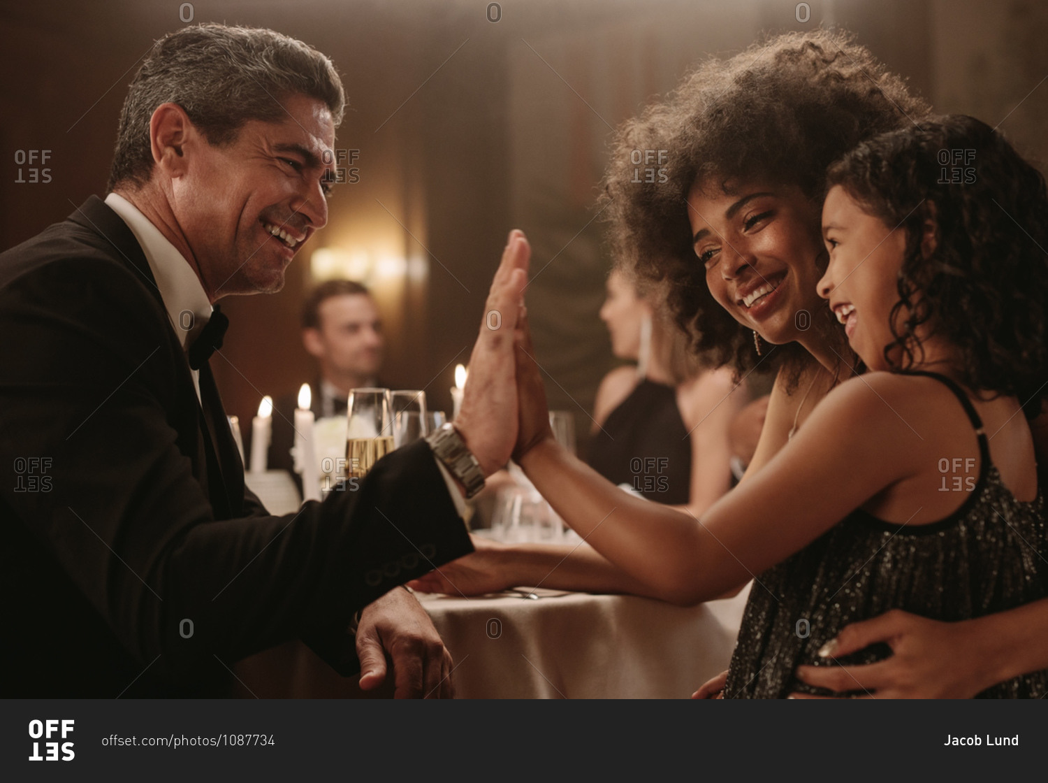 Mature man playing with a girl at dinner party with friends sitting around a dining table. Man giving high five to a little girl at gala night party.