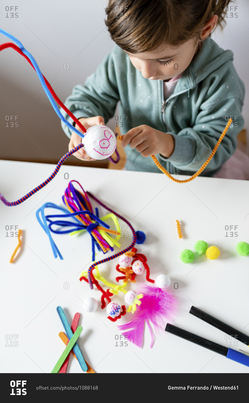 Cute girl making toys of styrofoam ball and pipe cleaners while sitting at table in living room