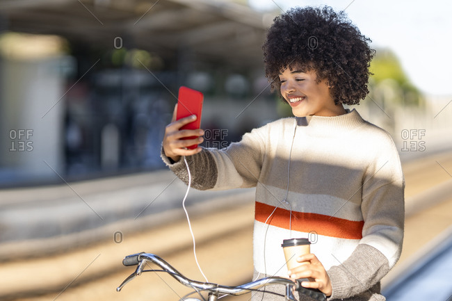 Young woman taking selfie while standing with bicycle and reusable cup at railroad station