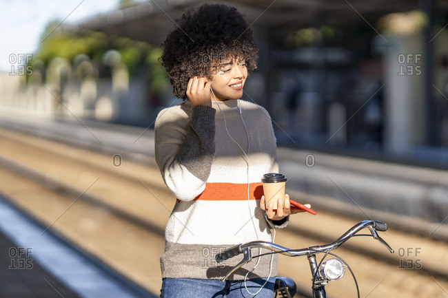 Smiling young woman with bicycle holding reusable cup while enjoying music at railroad station