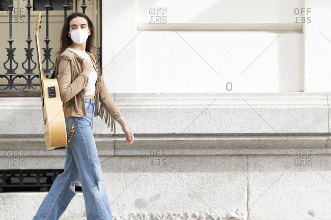 Female musician walking with guitar by building in city during pandemic