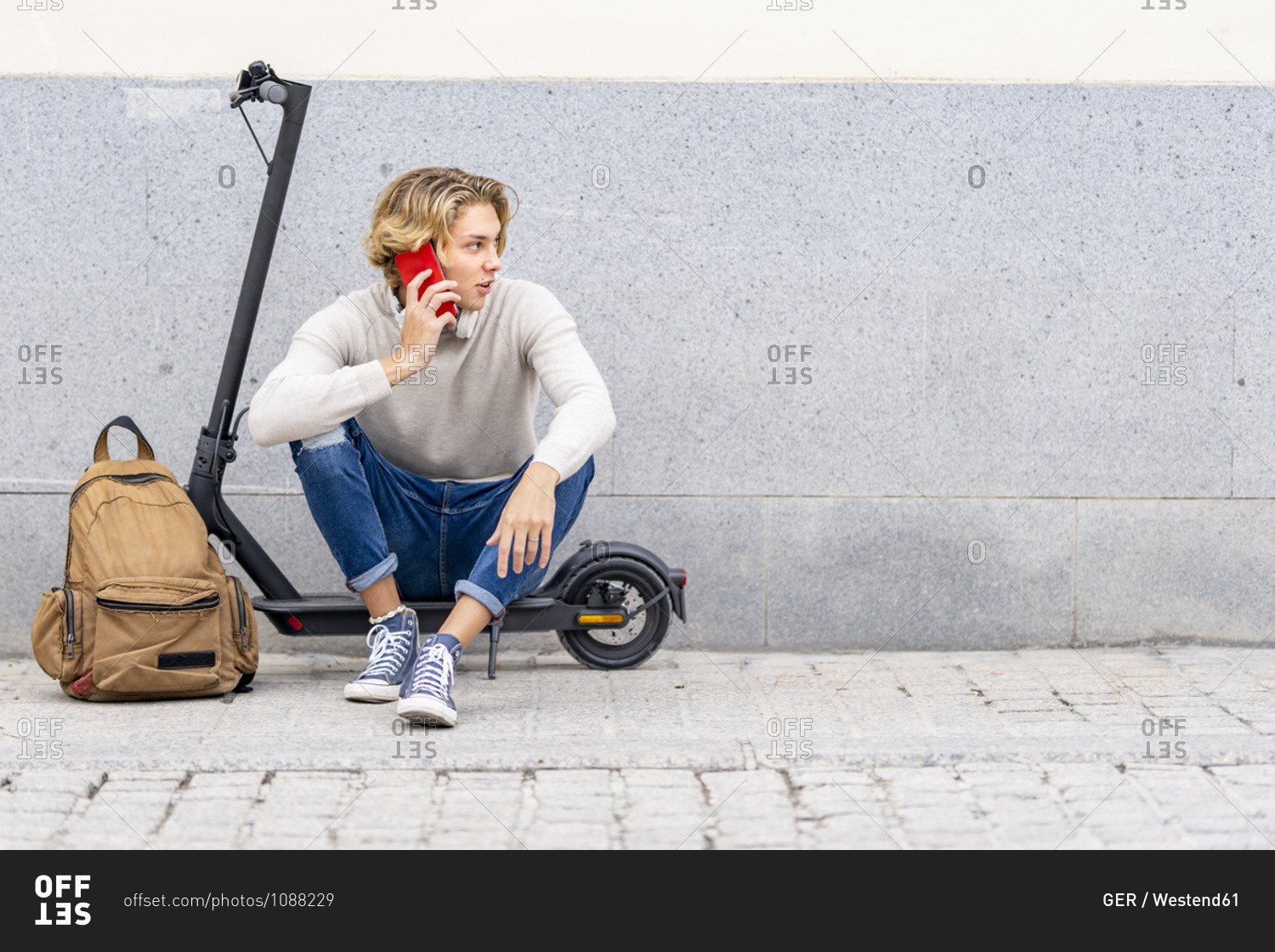 Man with backpack talking on mobile phone while sitting on electric push scooter against wall