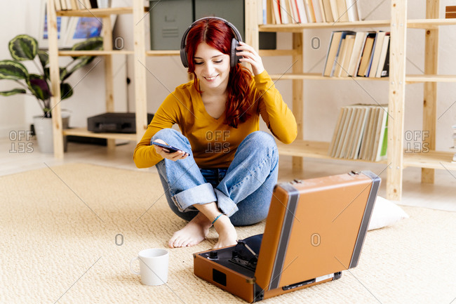 Smiling redhead woman wearing headphones using mobile phone while sitting by turntable at home