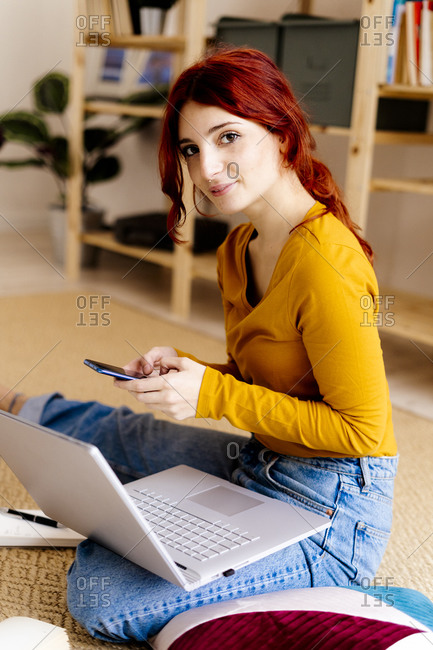 Redhead woman with laptop using mobile phone while sitting at home