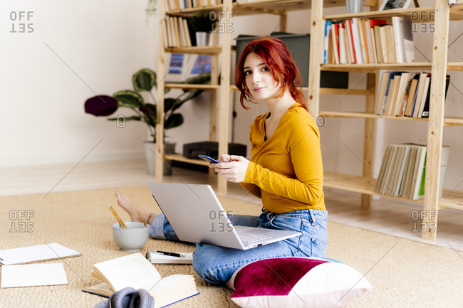 Young woman with laptop on lap using mobile phone while sitting at home