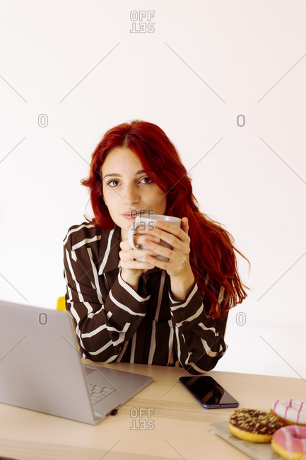 Redhead businesswoman drinking coffee while sitting by table at office